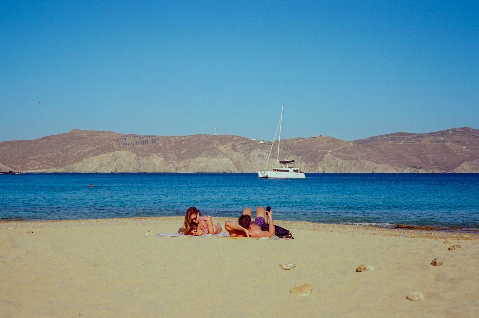 Two people sunbathing on a sandy beach with a sailboat.