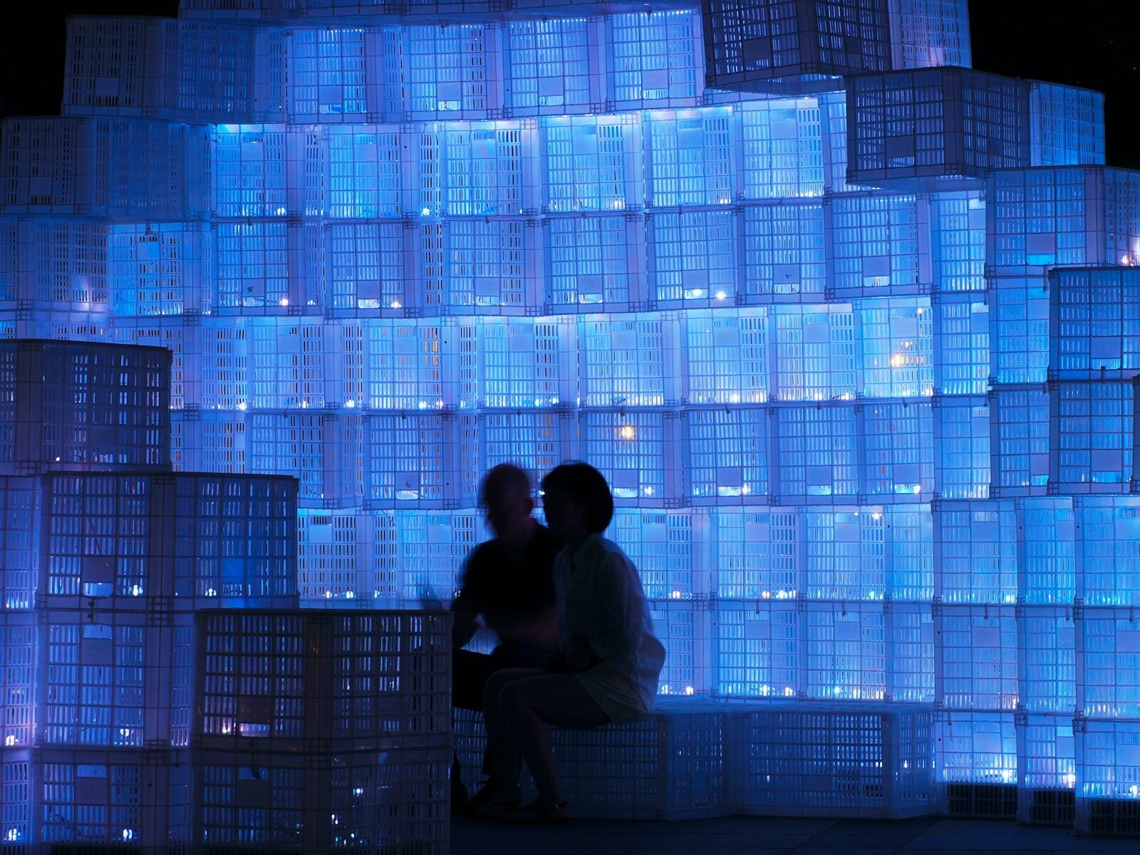 Silhouetted people sit by a wall of glowing blue boxes.